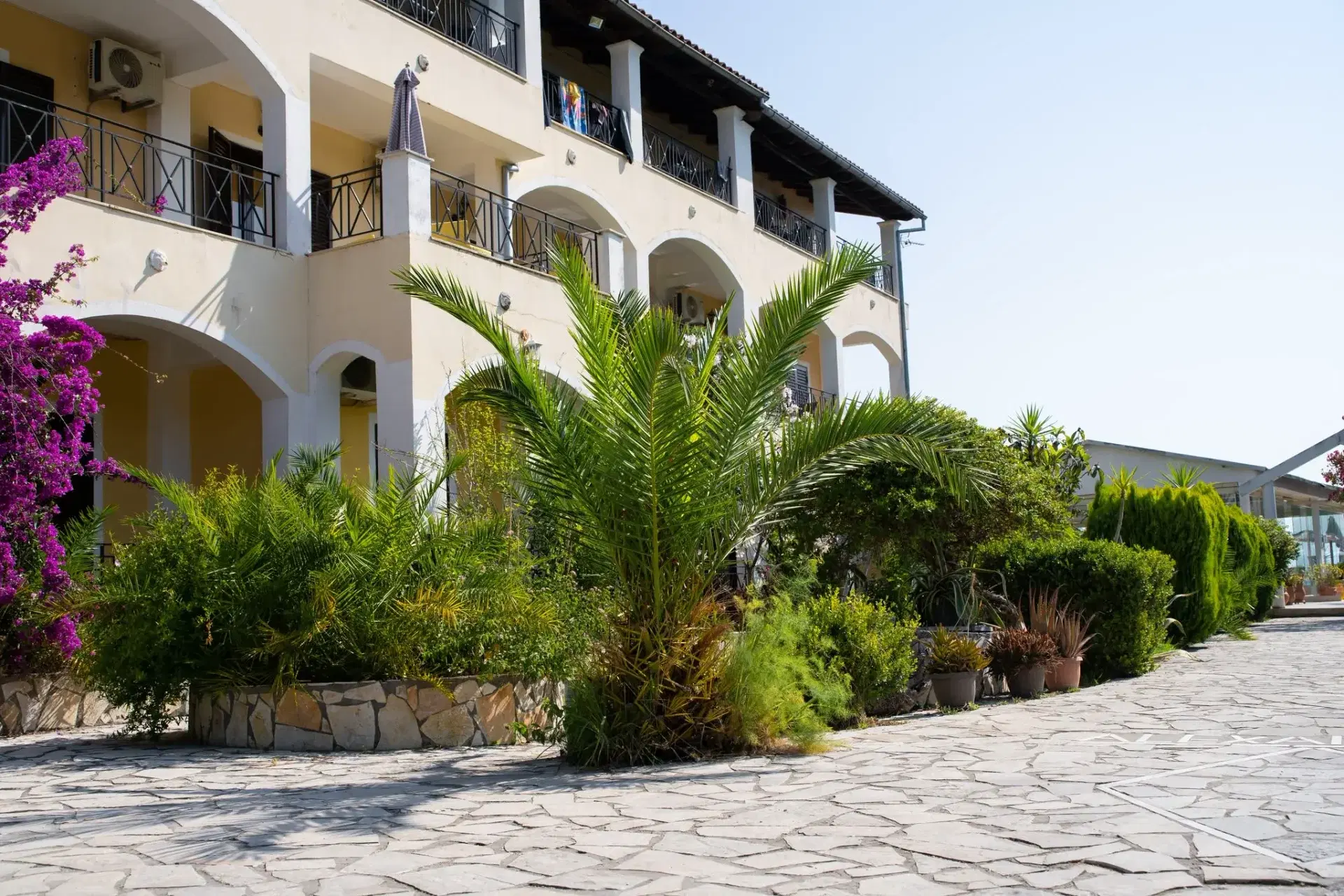 Az aparthotel a kerti sétányról nézve, erkélyeivel a pálmák és a bougainvillea mögött